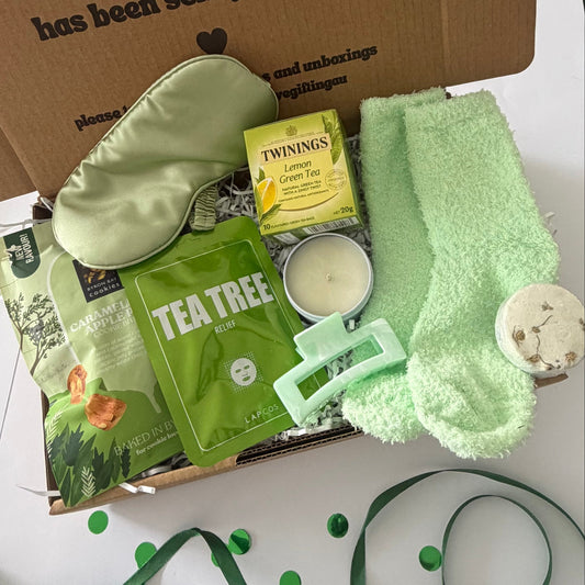 A curated gift box set containing a sage green silk eye mask, a fluffy sage green towel, a green tea tree face mask, a Twinings lemon tea packet, a small candle tin, and a bath bomb, arranged in a cardboard box with a green ribbon and confetti on a white surface.