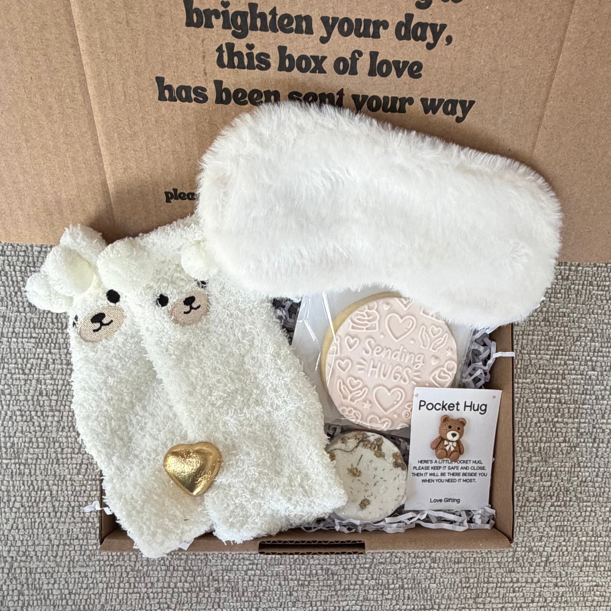 A cozy gift box set from Pocket Hug featuring a white plush eye mask, a white plush face mask with cute sheep faces and a gold heart, a round cream-colored soap, and a small card, all arranged inside an open brown cardboard box on a gray textured fabric background.
