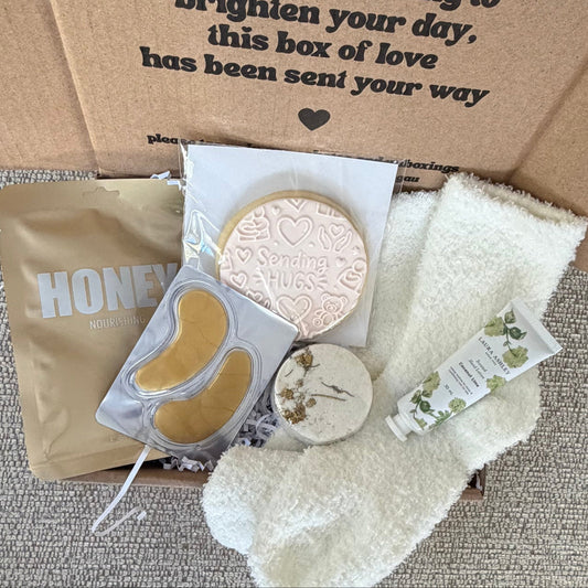 A curated  gift box opened honey lapcos mask, eye patches,  a small tube, and cream-colored socks. The kraft paper box has printed text reading 'a little something to brighten your day, this box of love has been sent your way'.