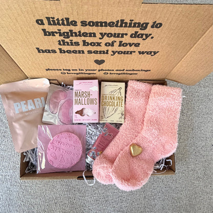 A curated gift box includes a pink plush sock with a gold heart, a pink bath bomb, a pink soap bar, a pink candle, and a pink pouch, all arranged with shredded paper inside a brown box printed with a message.