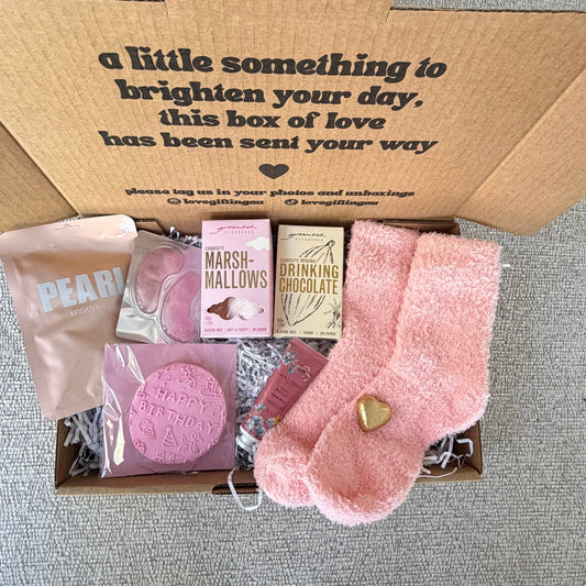 A curated gift box includes a pink plush sock with a gold heart, a pink bath bomb, a pink soap bar, a pink candle, and a pink pouch, all arranged with shredded paper inside a brown box printed with a message.