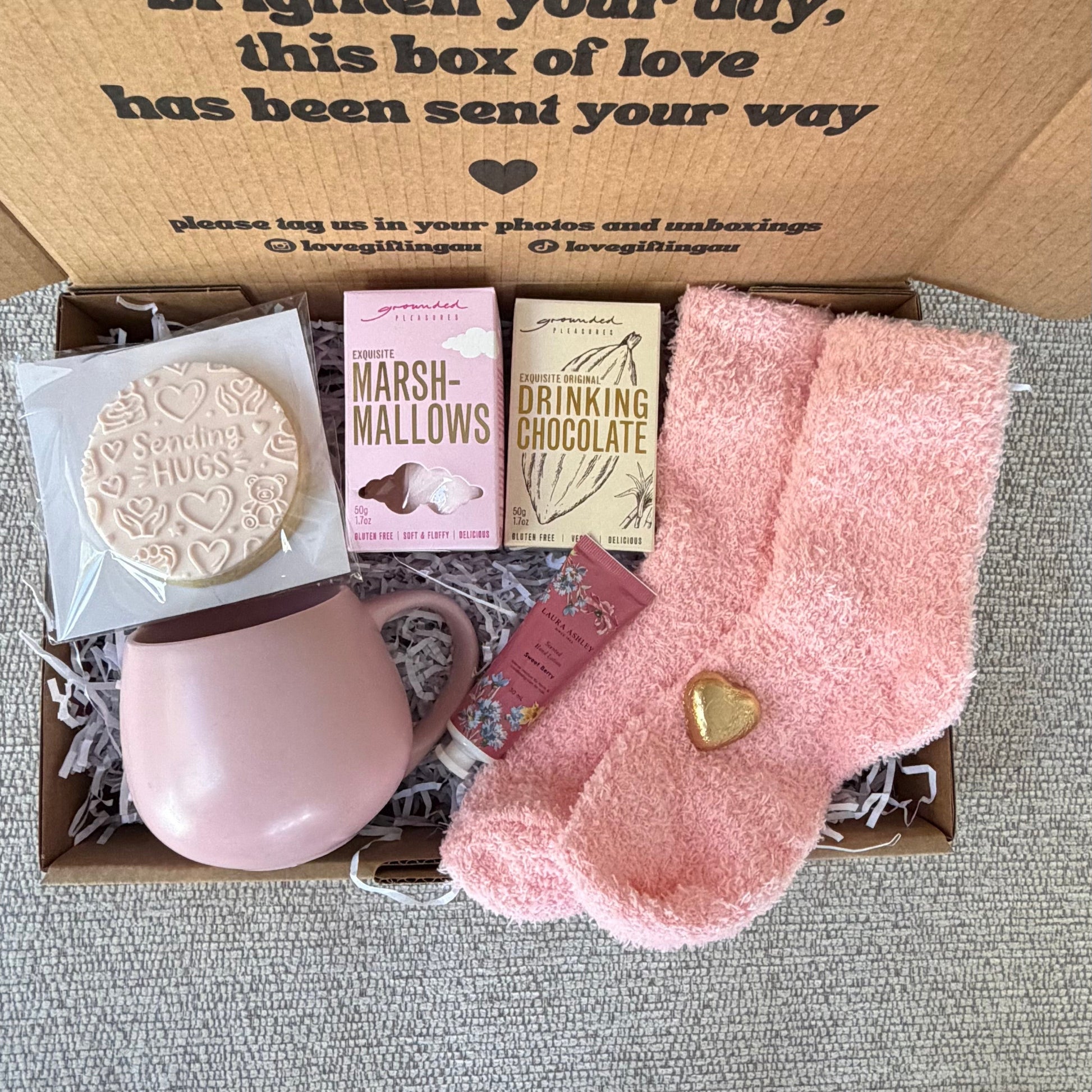 A cozy gift box containing a pink mug, a pink towel sock with a gold heart, a biscuit, and three chocolate boxes. The open cardboard box lid has printed text that reads 'a little something to brighten your day, this box of love has been sent i love you way'.
