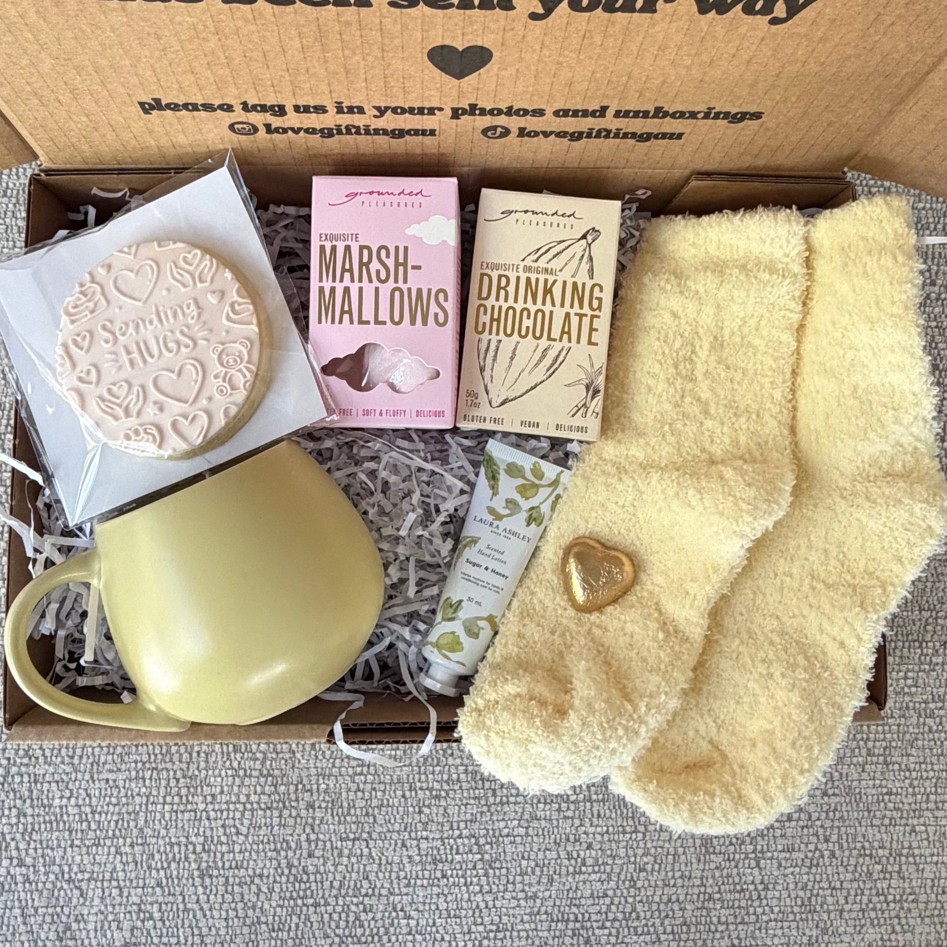 A curated gift box featuring a pale yellow ceramic mug, a round ] cookie, pink and beige chocolate heart, and a pair of fuzzy pale yellow socks, all arranged inside a brown cardboard box with a printed message.