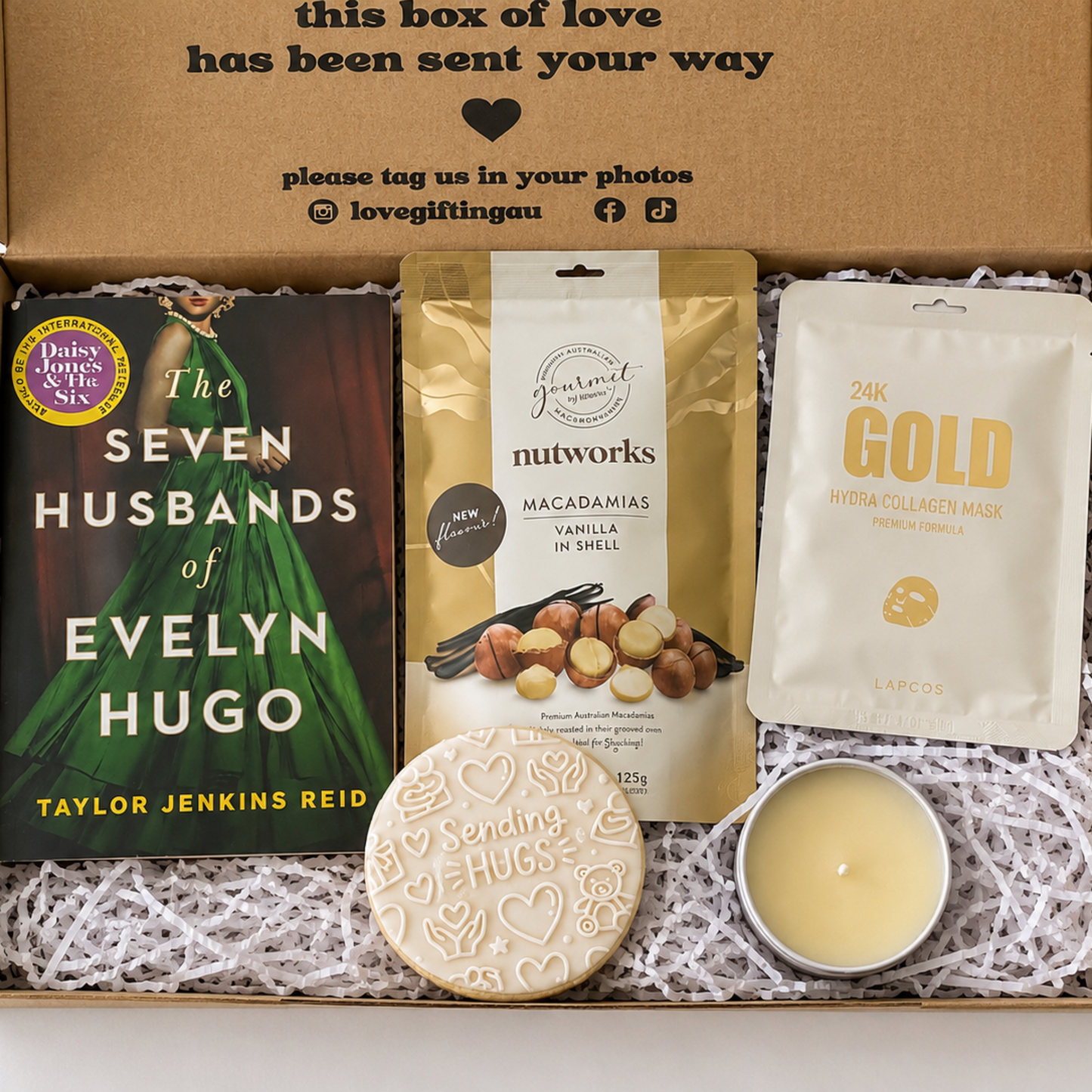 A curated gift box set displayed inside an open brown cardboard box with shredded paper filler. The set includes a book titled 'The Seven Husbands of Evelyn Hugo' by Taylor Jenkins Reid, a pouch of Nutworks Macadamia Flavour Gummies, a white pouch labeled '24K GOLD Facial Collagen Mask', a round heart-shaped cookie stamped with 'Sending Hug xHugs', and a small yellow candle.