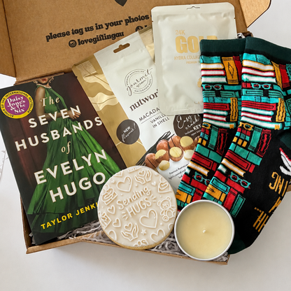 A curated gift box set opened on a white surface, containing a book titled 'The Seven Husbands of Evelyn Hugo,' a bag of MisoWor macadamia nut cookies, a white pouch with gold lettering, a colorful patterned pair of socks, a round embossed candle, and a small tea light candle. The open cardboard box has printed text at the top.