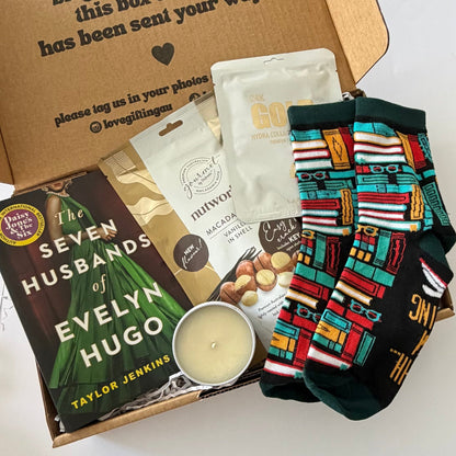 A curated gift box set in a brown cardboard box, featuring a book, a candle, packaged snacks, and a pair of colorful patterned socks on a white background.