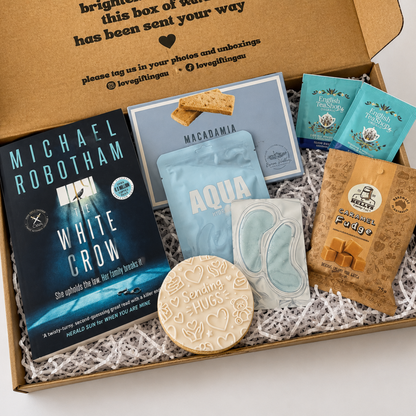 An open gift box filled with curated items on white shredded paper. Visible contents include a Michael Robotham book titled White Crow, a macadamia box, two blue tea bags, a brown paper snack bag, a light blue sachet, a small white item, and a round beige card that says 'Sending Hugs'.