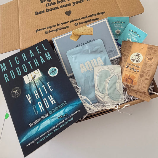 A curated gift box or subscription box filled with various items, including a Michael Robotham 'White Crow' book, a blue 'Aqua' face mask, a pair of white eye masks, a Macadamia Butter pouch, and other small packaged items, arranged in shredded paper inside an open cardboard box.