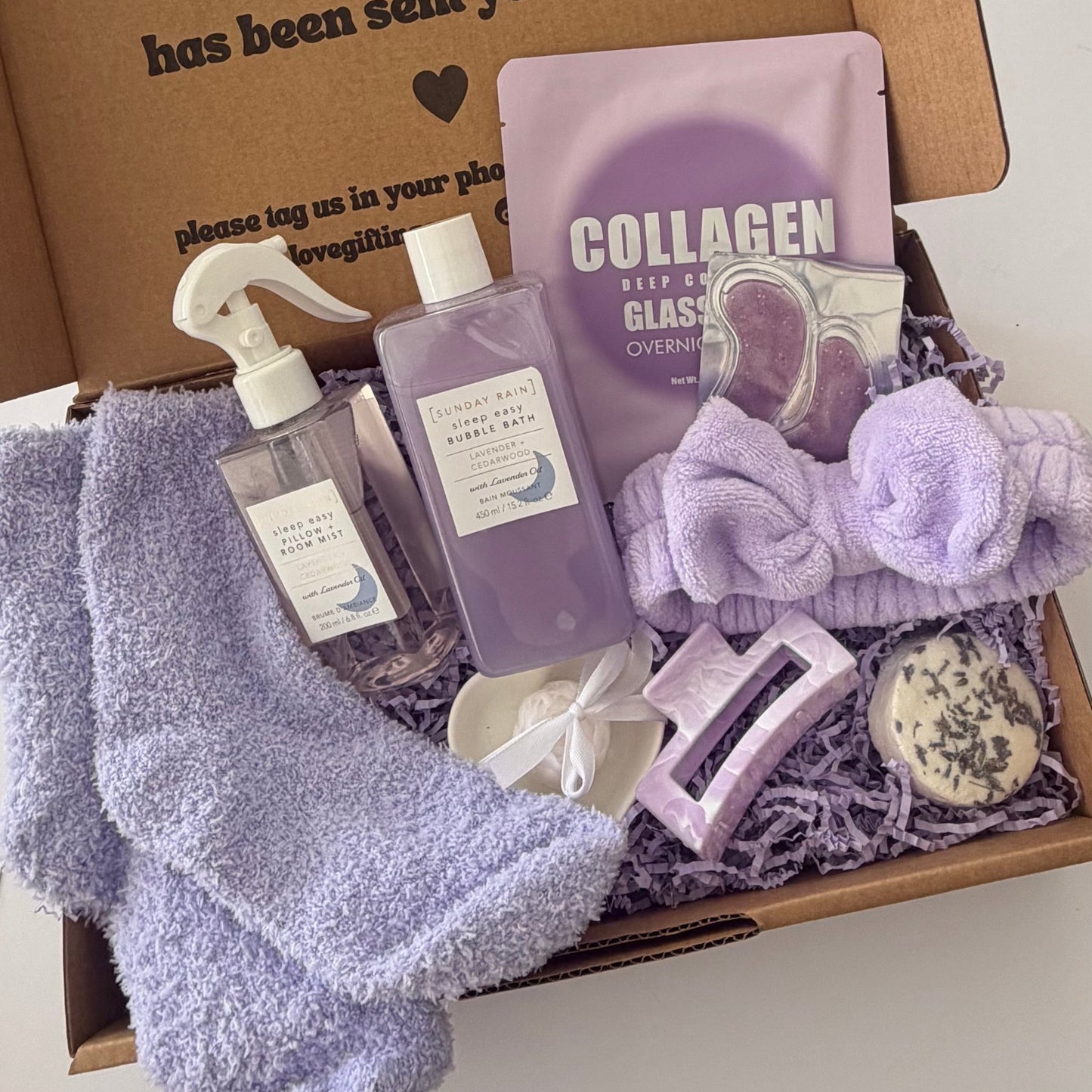 A curated self-care gift box filled with lavender and purple items, including a plush towel, a spray bottle, a collagen sheet mask packet, a headband, a small soap bar, and a bar of soap, arranged in a cardboard box with shredded paper filler.