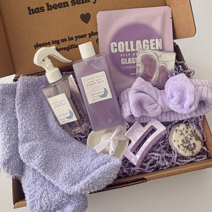 A curated self-care gift box filled with lavender and purple items, including a plush towel, a spray bottle, a collagen sheet mask packet, a headband, a small soap bar, and a bar of soap, arranged in a cardboard box with shredded paper filler.