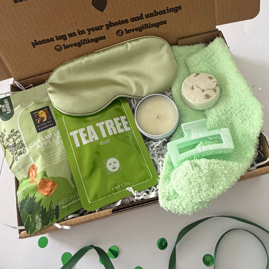 A curated gift box set containing a sage green satin sleep mask, a green tea tree face mask packet, a small candle, a bath bomb, a green plush towel, and a green soap bar, arranged inside an open cardboard box with a green ribbon in the foreground.