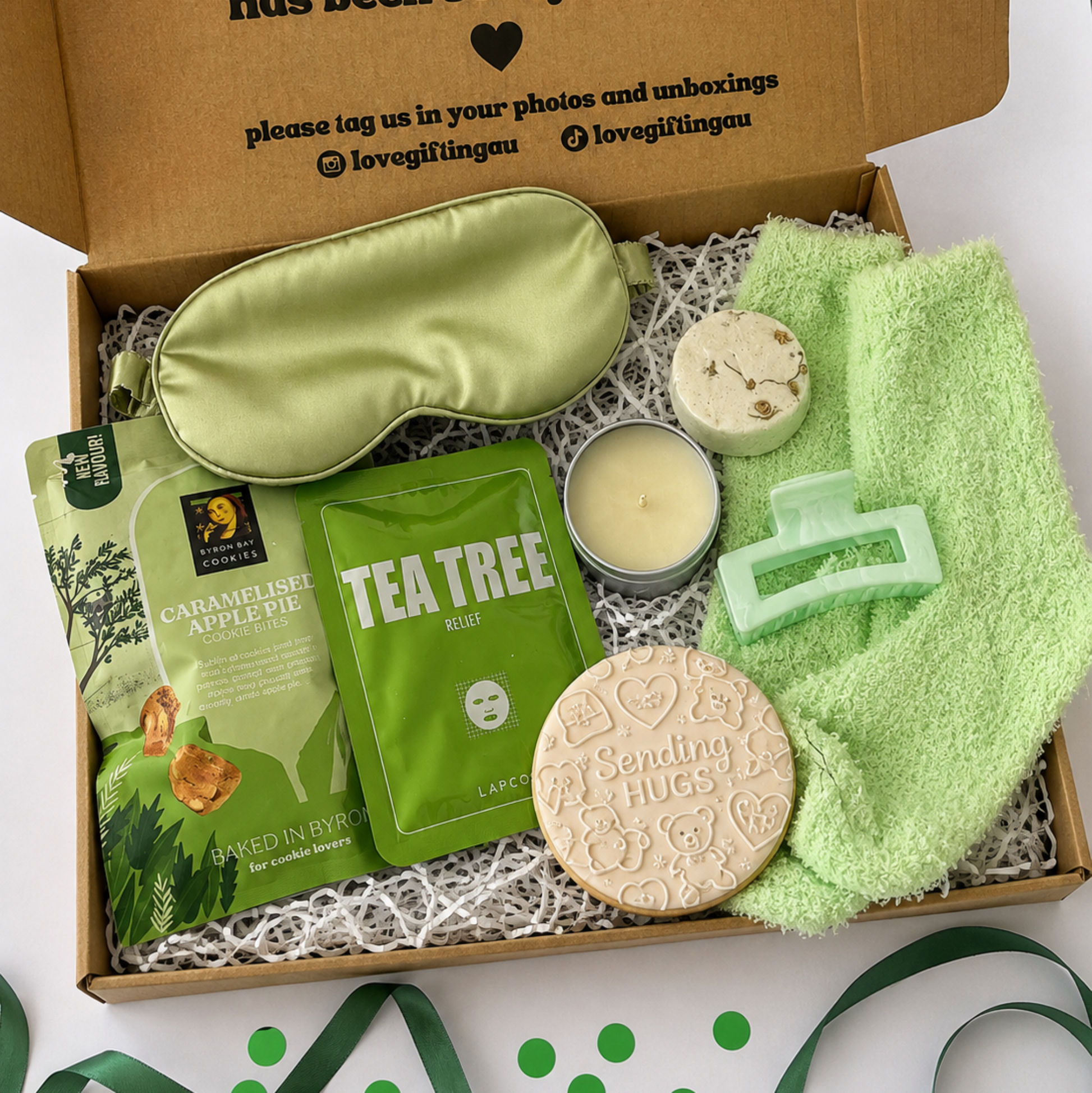 A curated gift box set in a brown cardboard box with shredded paper filler, featuring a light green satin sleep mask, a green tea tree mask packet, a green apple tea packet, a small candle, a green hair clip, a round beige soap bar, and a green towel, arranged on a white surface with green ribbon accents.