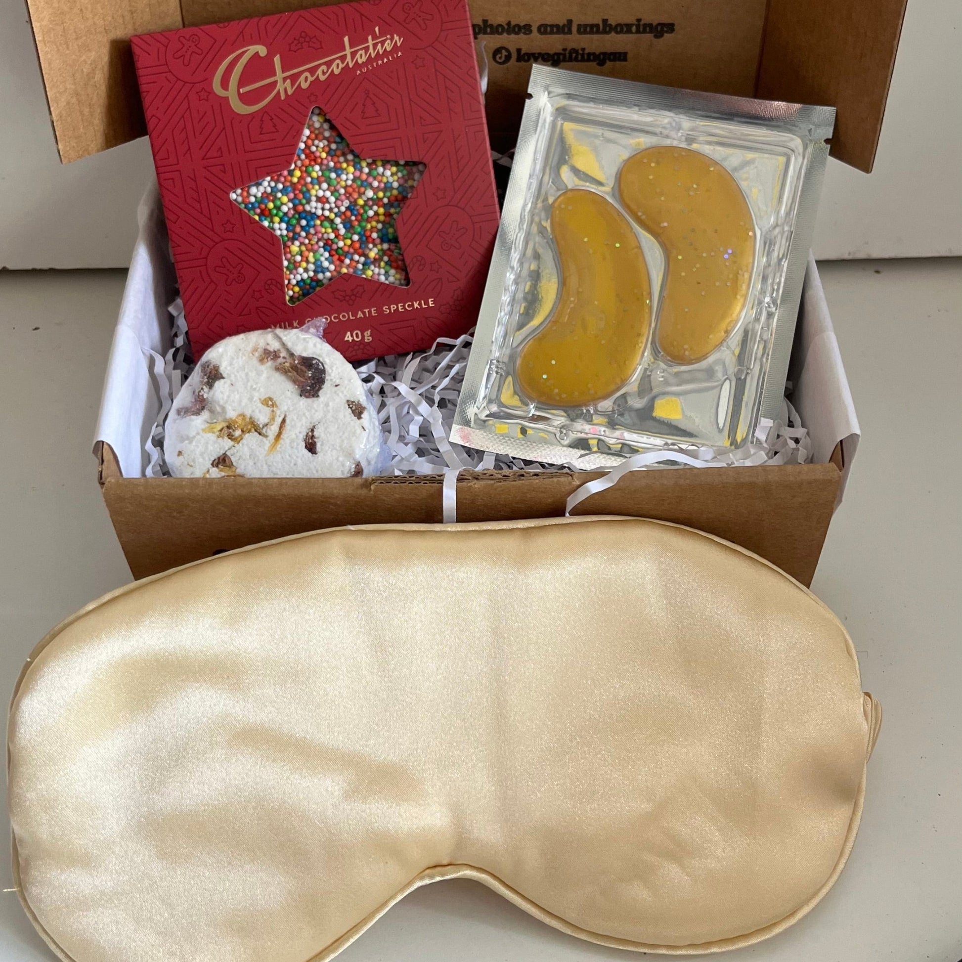 Box of self-care items including a gold sleep mask, star-shaped chocolate , shower steamer and gold under eye masks
