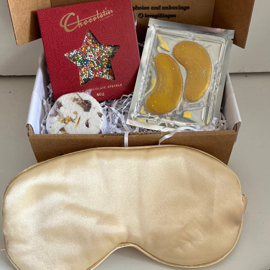 Box of self-care items including a gold sleep mask, star-shaped chocolate , shower steamer and gold under eye masks