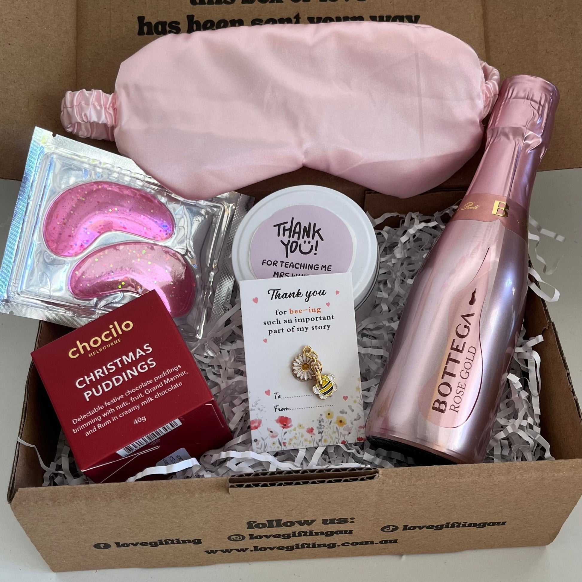 Gift box with pink sleep mask, chocolate, and bottle of champagne on a white background