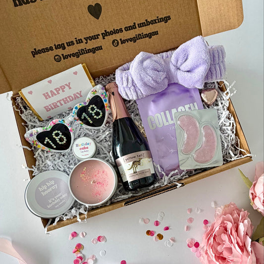 A curated birthday gift box filled with spa and self-care items, photographed from above on a white surface with pink flowers around it. The open kraft box lid has printed text and social handles. Prosecco , happy birthday chocoalte bar, 18th glasses, candle, lapcos facemask, under eye, towelling headband, birthday cake lipbalm, chocolate heart.