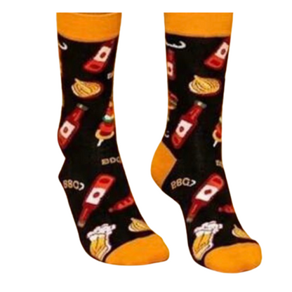 A pair of crew socks with a black background and a colorful pattern of hot dogs, beer mugs, and the word 'BBQ'. The socks have bright orange toes, heels, and cuffs, and are shown against a plain white background.