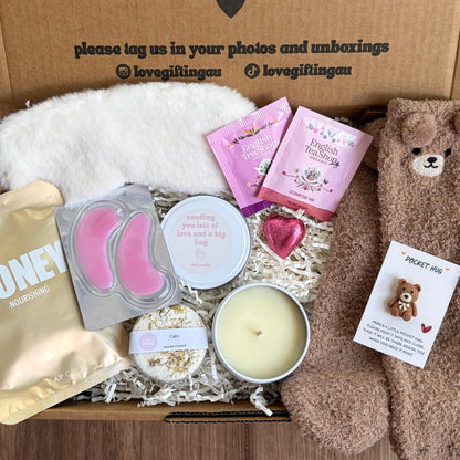 Greeting card with 'a little something to brighten your day' message, surrounded by a box of products including a candle, face masks, and a teddy bear.