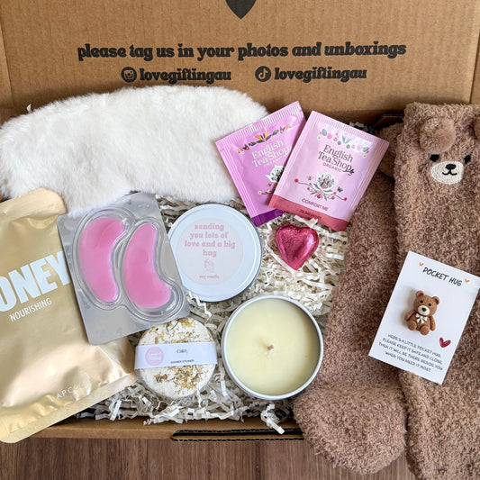 Greeting card with 'a little something to brighten your day' message, surrounded by a box of products including a candle, face masks, and a teddy bear.