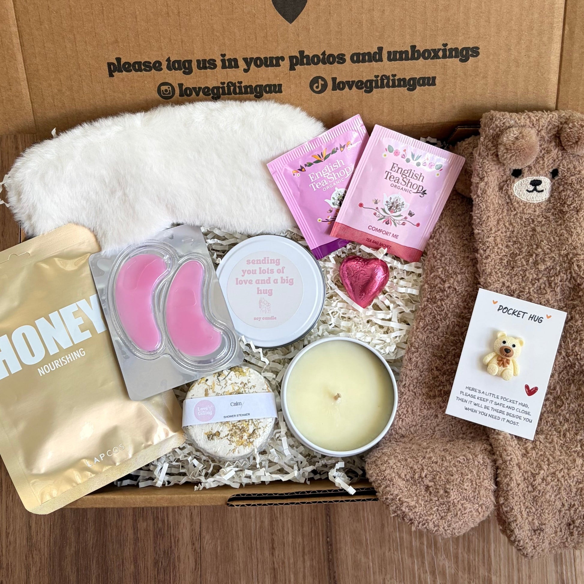 Greeting card with 'a little something to brighten your day' message, teddy bear, skincare products, and a candle on a wooden surface.