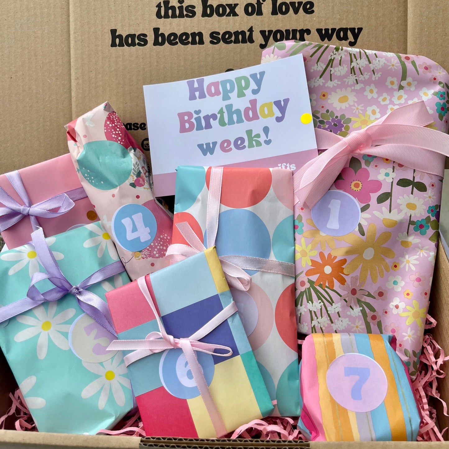 Box of colorful birthday-themed gift boxes with a card inside, on a cardboard background.
