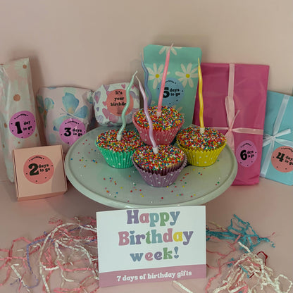 Decorative birthday setup with colorful cupcakes, gift boxes, and a 'Happy Birthday week!' card on a light pink background.