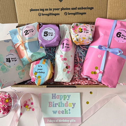 A curated birthday gift box set displayed on a white surface, featuring a brown cardboard box with printed text, wrapped birthday treats, colorful gift tags, a pink wrapped present with a blue ribbon, a small jar of pink candies, and a pastel 'Happy Birthday week!' card.