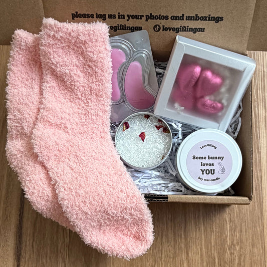 Box of self-care items including pink socks, candles, and a heart-shaped object on a wooden surface.