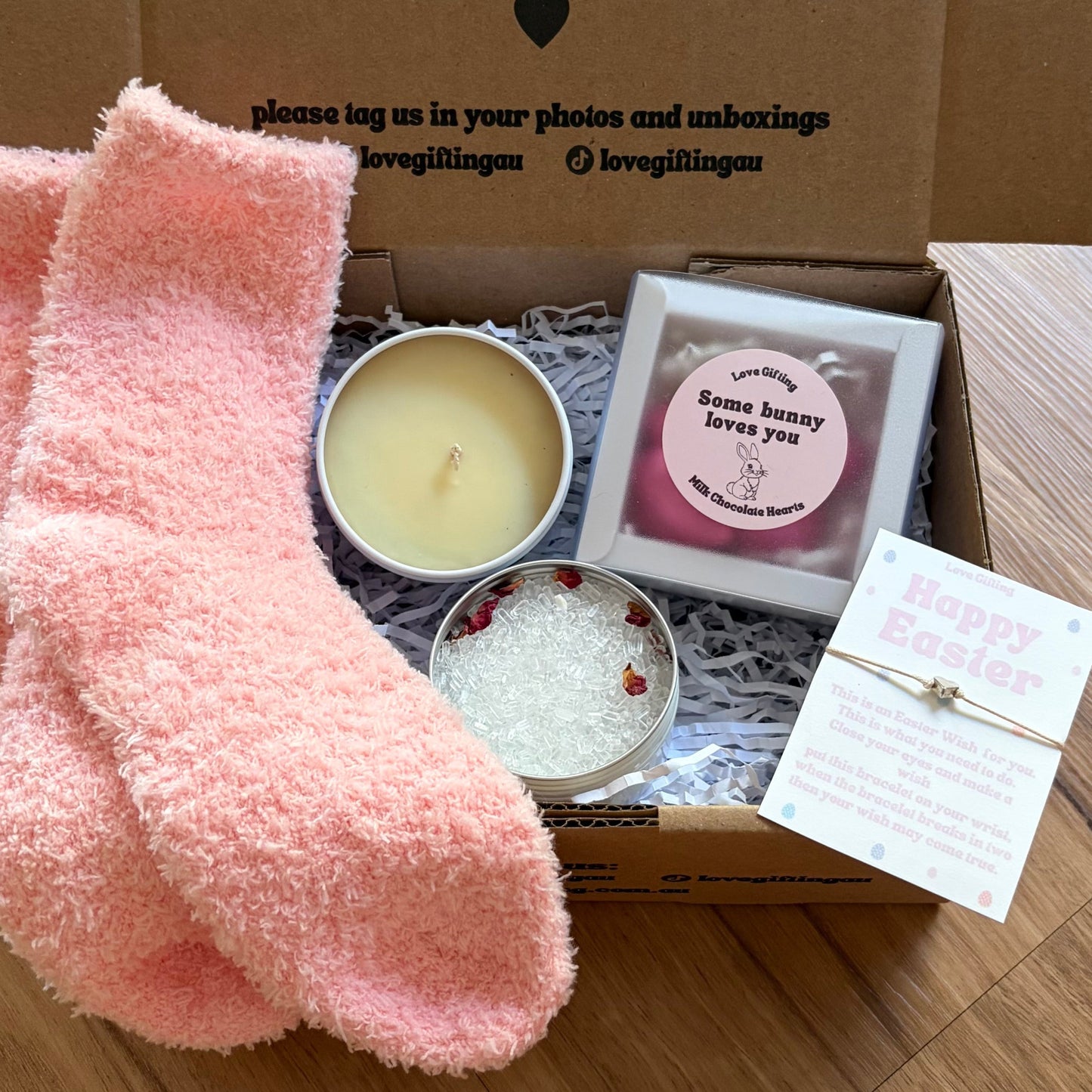 Box of self-care items including pink socks, a candle, and a card on a wooden surface.