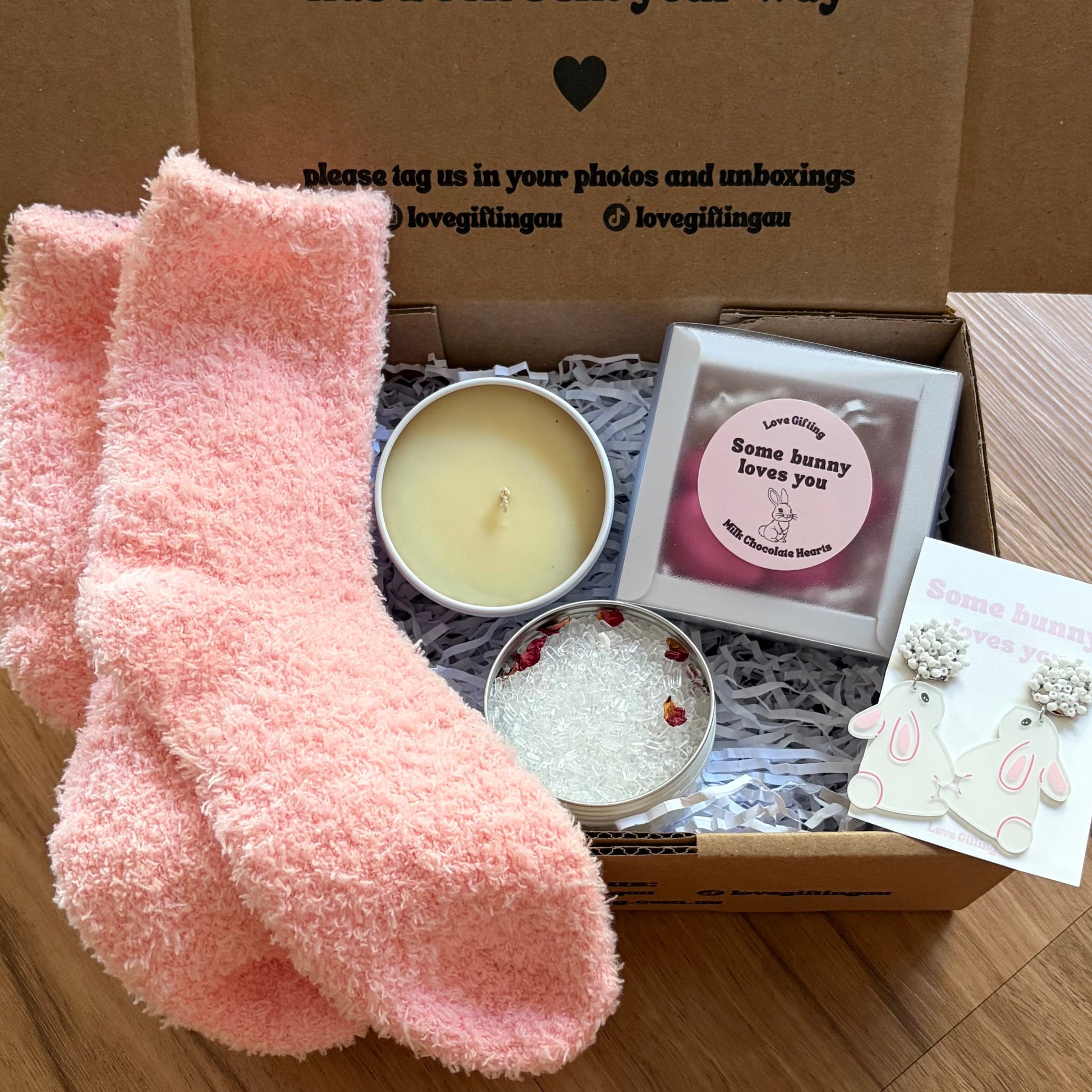 Box of self-care items including pink socks, a candle, and other small products on a wooden surface.