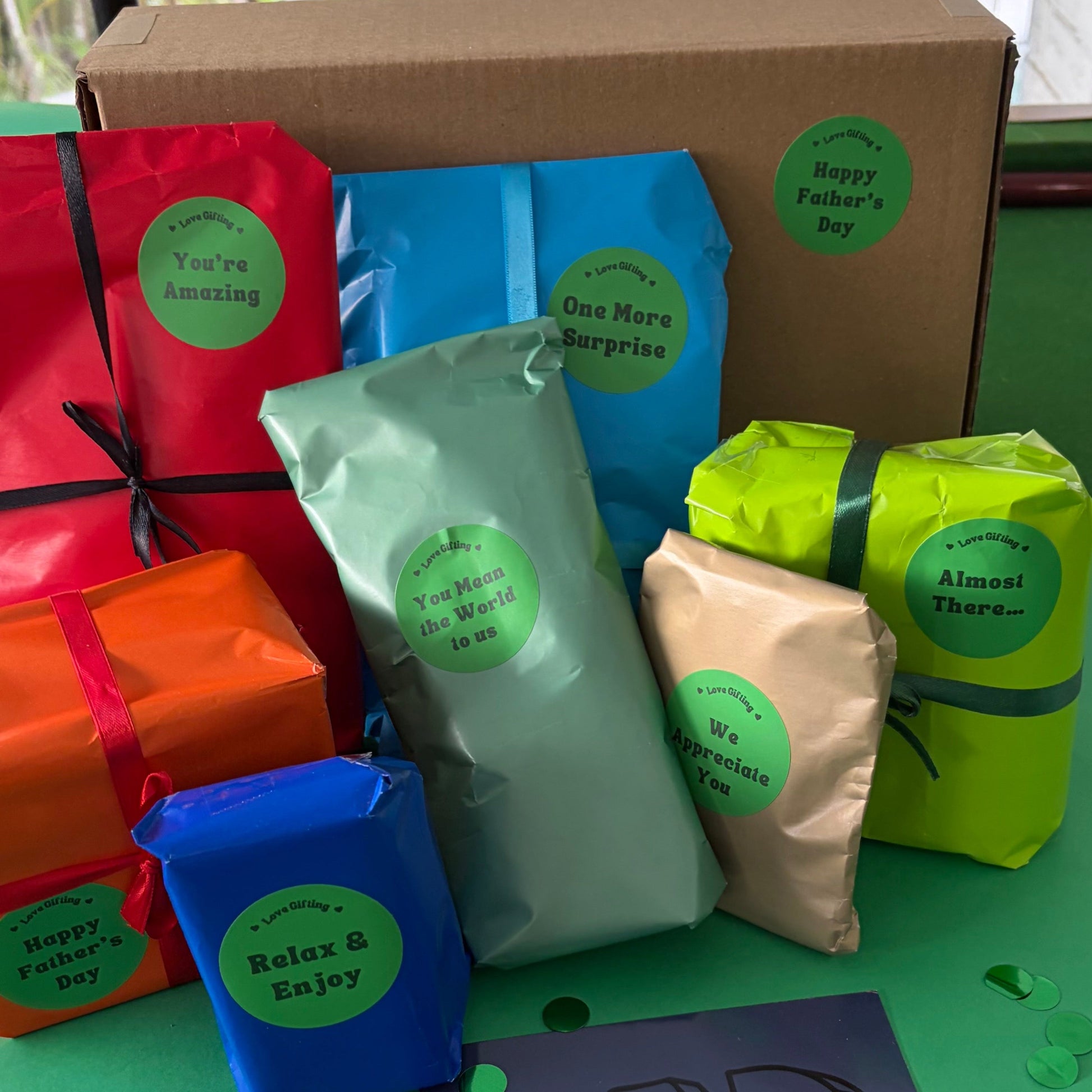 A curated gift set for Dad displayed on a green table, featuring a large brown cardboard box, several colorful gift bags in red, blue, mint green, lime green, and beige, a blue gift box, and a purple card that says 'DAD' with green confetti scattered around.