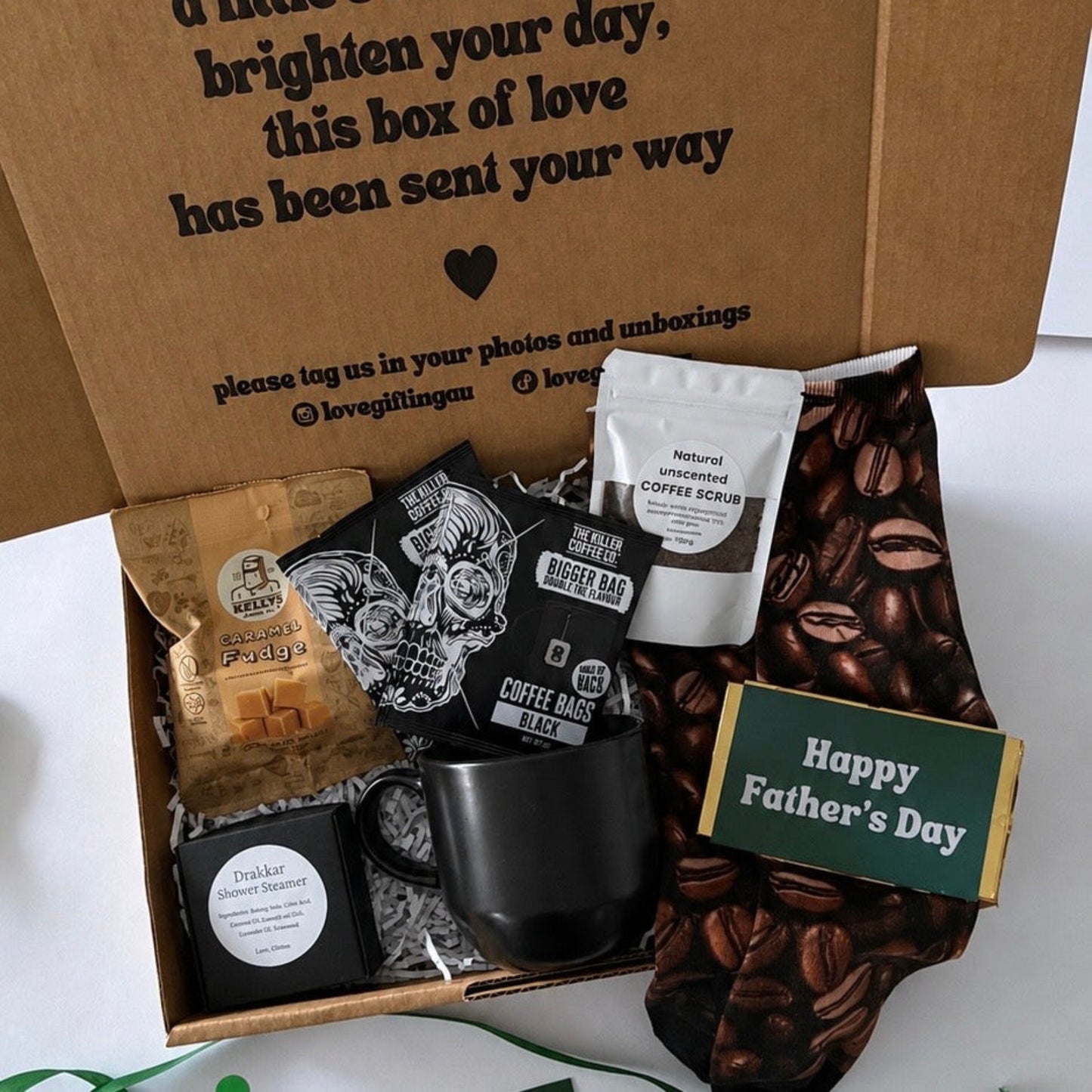 A Father's Day gift box filled with coffee-themed items, including a black coffee mug, a bag of coffee beans, a pouch of coffee scrub, a small coffee grinder, and a 'Happy Father's Day' card, set in a brown cardboard box with a printed message.