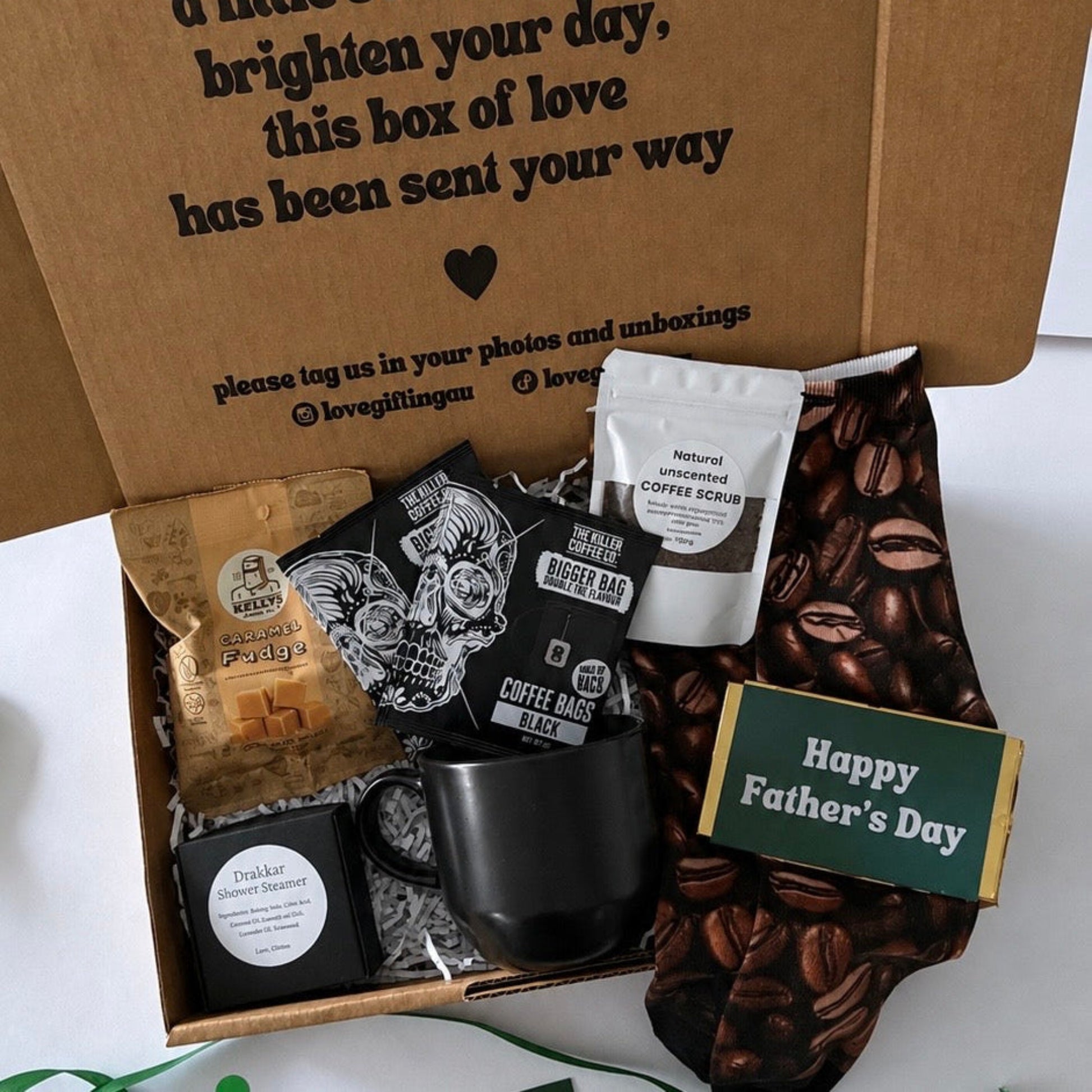 A Father's Day gift box filled with coffee-themed items, including a black coffee mug, a bag of coffee beans, a pouch of coffee scrub, a small coffee grinder, and a 'Happy Father's Day' card, set in a brown cardboard box with a printed message.