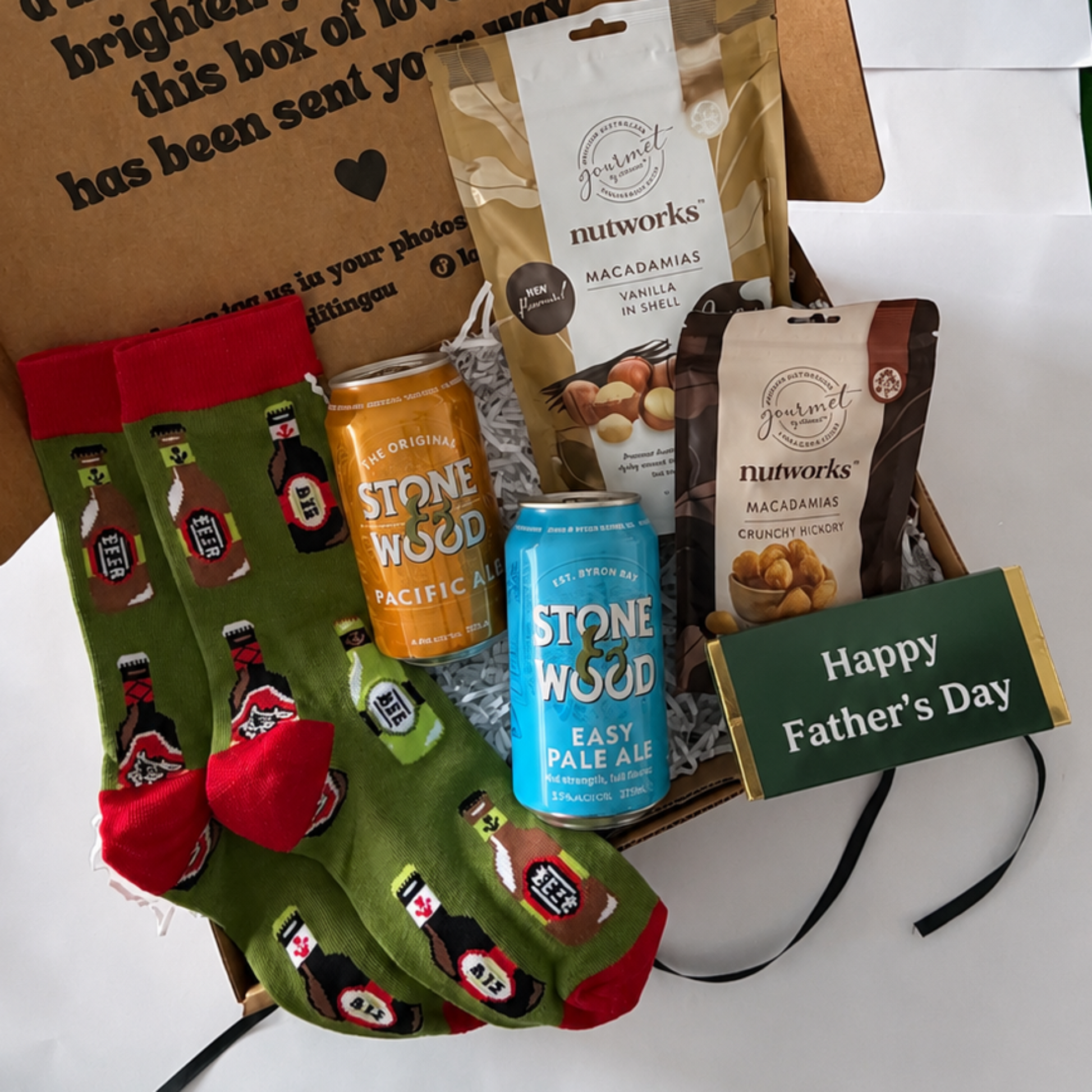 A curated Father's Day gift box containing a brown kraft envelope with printed text, green novelty socks with red accents, a can of Stone Wood Coffee, a bag of Nutworks almonds, and a green 'Happy Father's Day' card, arranged on white paper over a green surface.