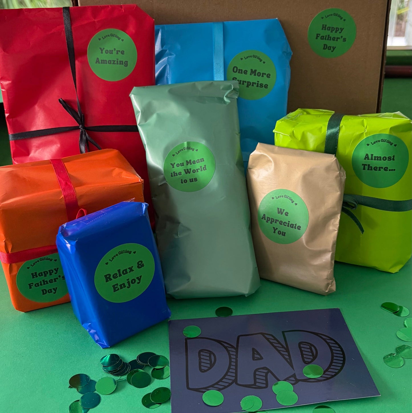 A Father's Day gift set featuring multiple packaged tea bags in colorful wrapping paper and pouches, arranged on a green surface with a cardboard box in the background. The packages are wrapped in red, blue, orange, sage green, beige, and lime green paper with green circular labels.