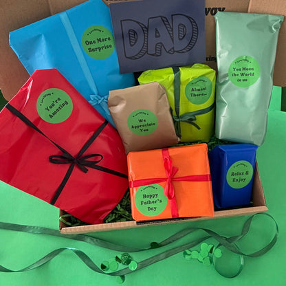 A Father's Day gift box filled with colorful packaged treats and surprises, including a red gift bag, blue pouches, a green bag, a brown paper bag, an orange wrapped box, and a small blue box, arranged in a cardboard box with a green surface underneath.