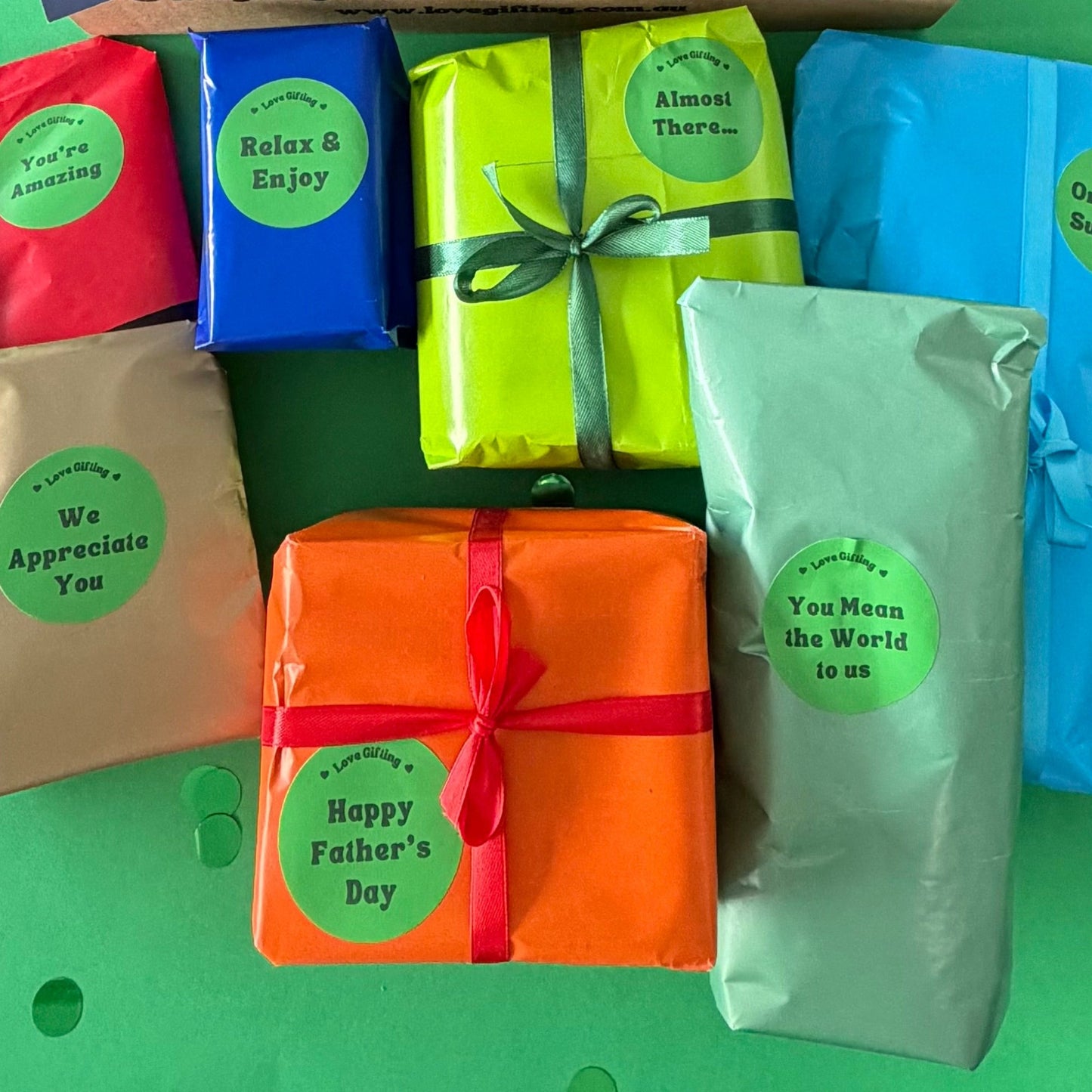 A Father's Day gift set featuring a brown cardboard box labeled 'Love Gifting' with a blue 'DAD' card, surrounded by six wrapped gift boxes in red, beige, blue, green, orange, and mint with green circular stickers reading 'Happy Father's Day' and other phrases.