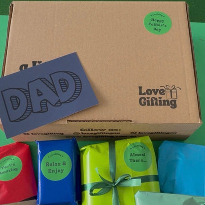 A cardboard gift box labeled 'Love Gifting' with a green 'Happy Father's Day' sticker, alongside a blue card reading 'DAD' and several wrapped presents in red, blue, lime green, and light blue on a green surface.