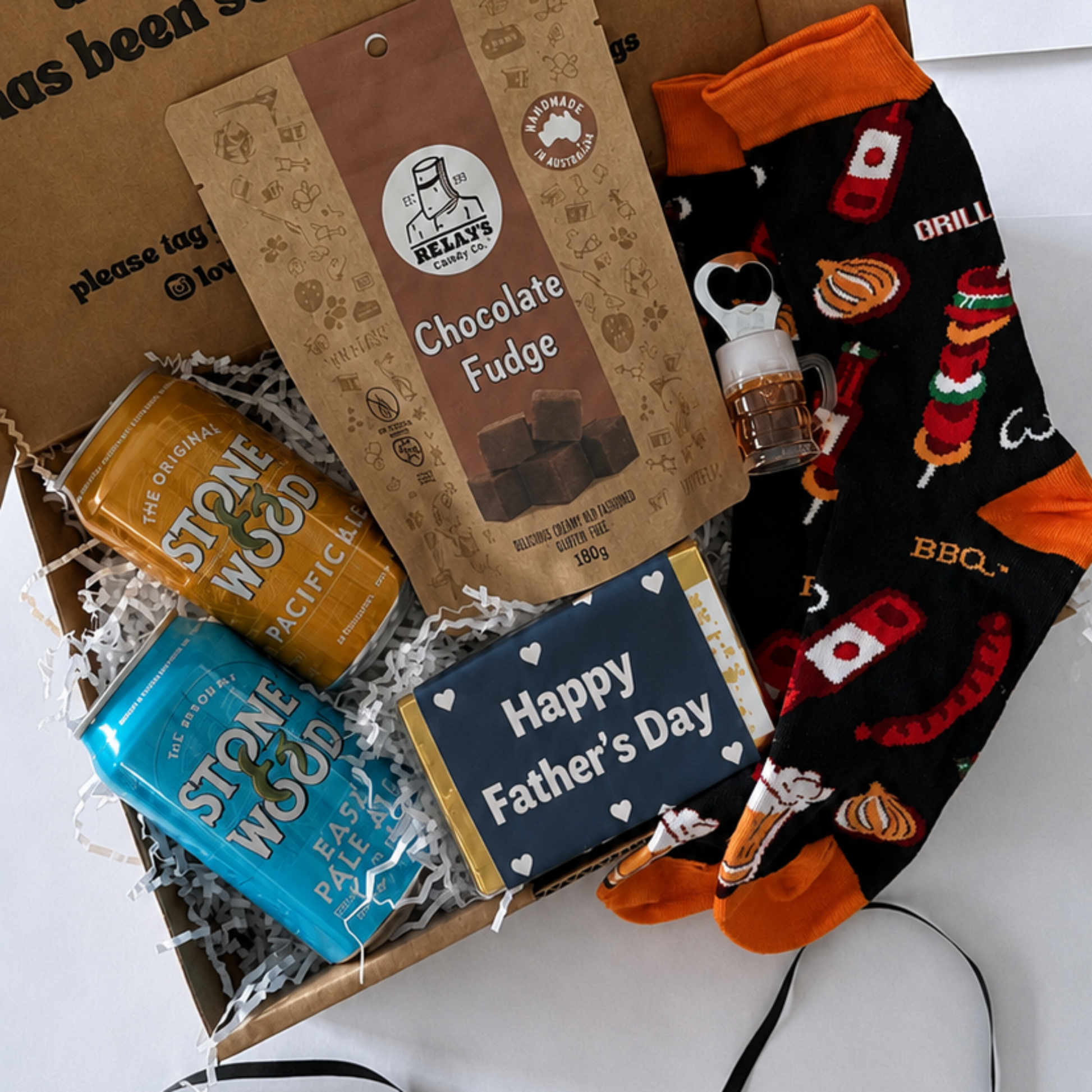 A Father's Day gift box set containing a chocolate fudge bar, a bottle of stone wood beer and a pair of black and orange socks with a campfire design, all arranged inside an open kraft paper box with a printed message.