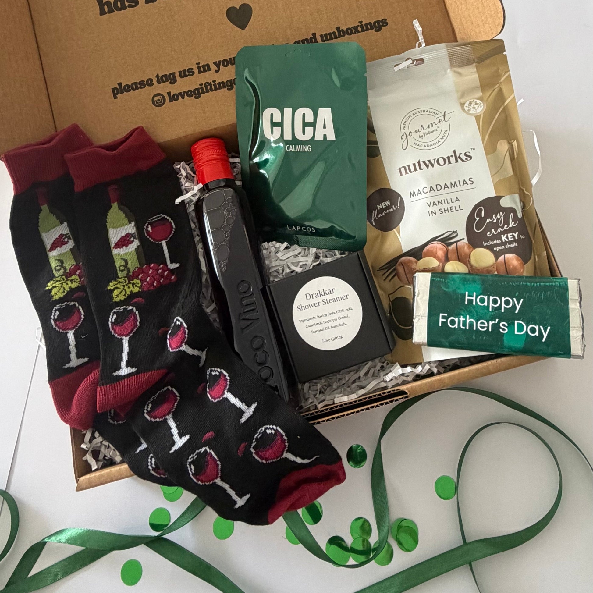 A curated Father's Day gift box filled with themed items: black crew socks with a wine bottle and glass pattern, a dark bottle with a red cap, a green bag of Cica Flavors protein powder, a bag of Nutworks macadamia vanilla energy bars, and a green 'Happy Father's Day' card, arranged on a white surface with green ribbon and confetti.