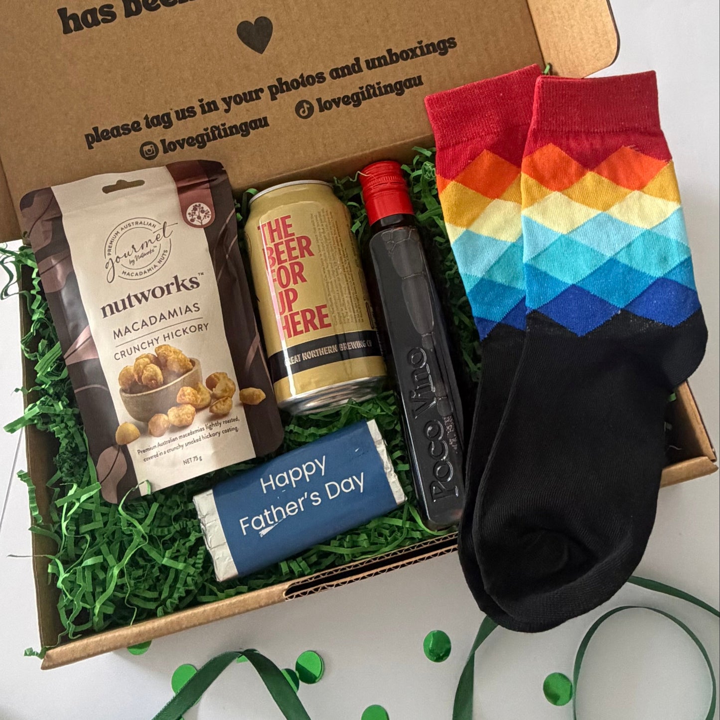 A Father's Day gift box filled with treats and a pair of rainbow socks. The box contains a bag of Nutworks macadamia chocolate milk chocolate, a can of 'The Beer For Dri-Fiere', a black bottle of Grey Goose bourbon, and a blue bar of soap that says 'Happy Father's Day'.
