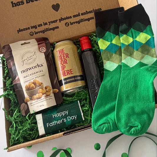 A curated Father's Day gift box containing a bag of Nutworks macadamia chips, a can of beer, a bottle of dark liquor, and a green 'Happy Father's Day' card, arranged in green shredded paper inside a cardboard box next to a pair of bright green argyle socks.