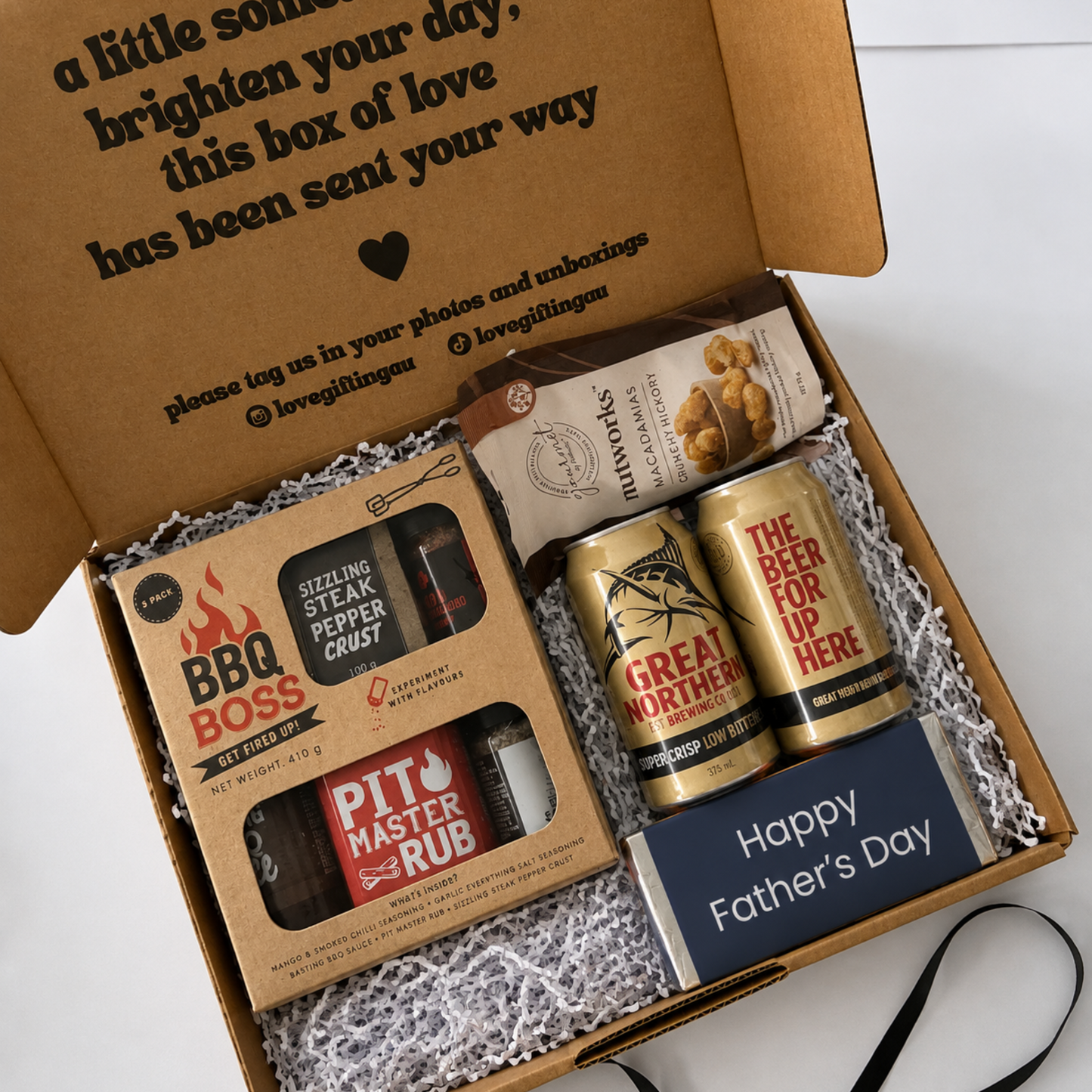 A curated Father's Day gift box set in a brown cardboard box with white crinkle paper filler. The set includes a BBQ Boss rub and pepper grinder, a Great Northern beer can, a Snickers 'The Beer For Up Here' can, a bag of mac and cheese, and a navy blue card reading 'Happy Father's Day'.