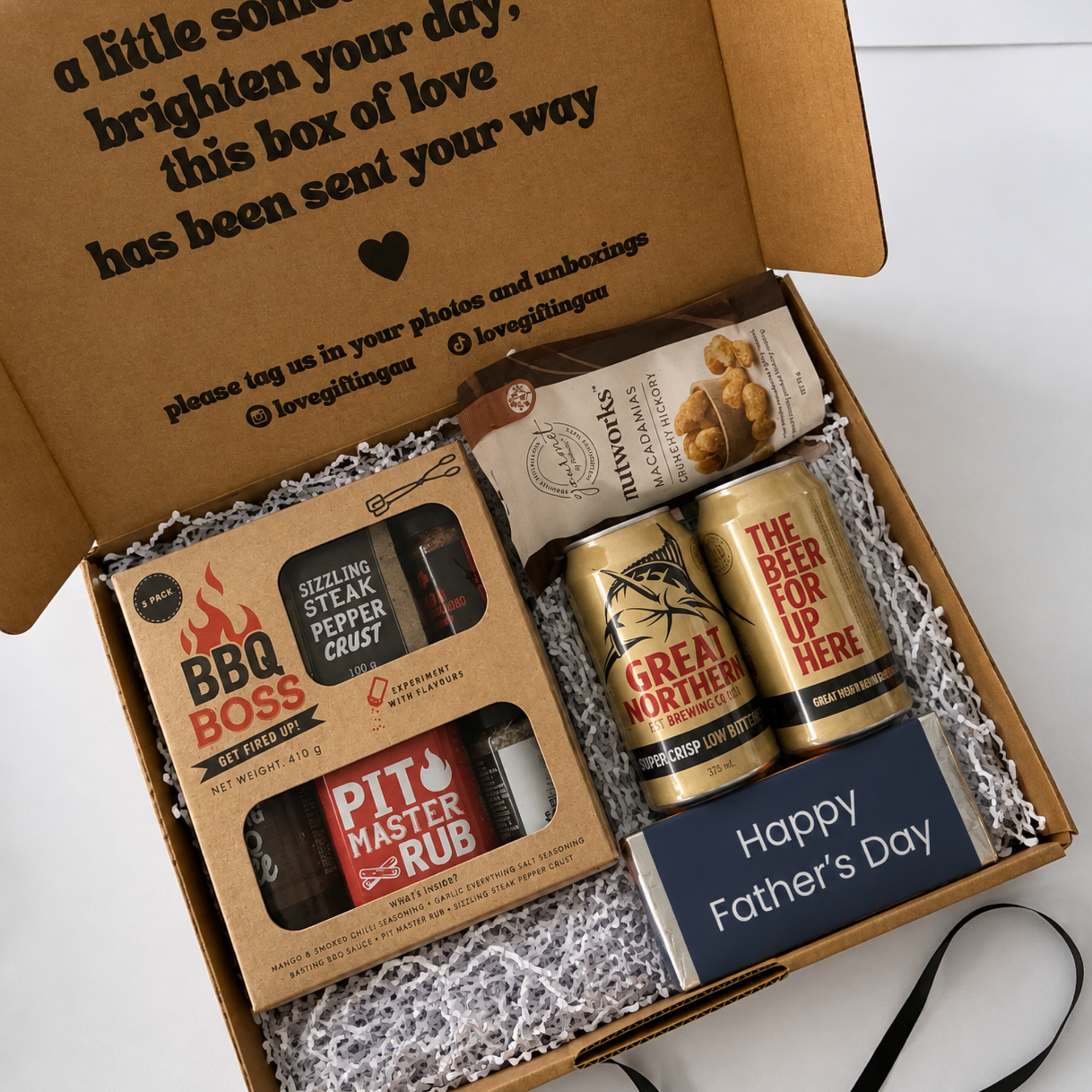 A curated Father's Day gift box set in a brown cardboard box with white crinkle paper filler. The set includes a BBQ Boss rub and pepper grinder, a Great Northern beer can, a Snickers 'The Beer For Up Here' can, a bag of mac and cheese, and a navy blue card reading 'Happy Father's Day'.