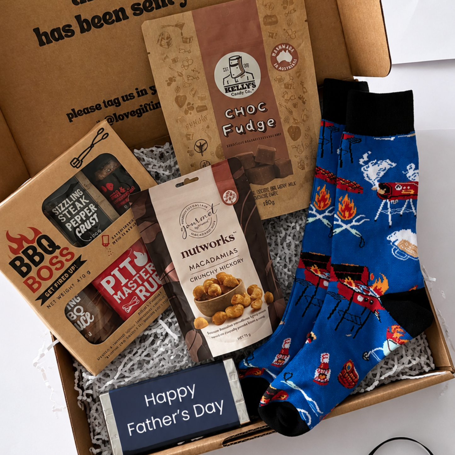A Father's Day gift box filled with treats and socks, photographed from above on a white surface. The open kraft box contains a BBQ Boss rub, Pithworks caramel chips, a chocolate fudge box, and a pair of blue crew socks with a cowboy motif.