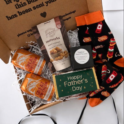A Father's Day gift box filled with treats and socks on a white background. The box contains two orange cans of Great Value canned soda, a bag of Nutworks macadamia, a small black box, a green 'Happy Father's Day' card, and a pair of black socks with orange cuff trim and a red and white BBQ pepperoni pattern.