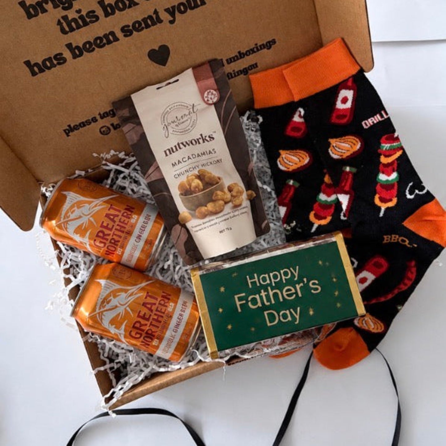 A Father's Day gift box set on a white background, containing a bag of Nutwags Macadamia nuts, two cans of Great Value orange soda, a small green 'Happy Father's Day' card, and a pair of black and orange socks with a barbecue grill pattern.