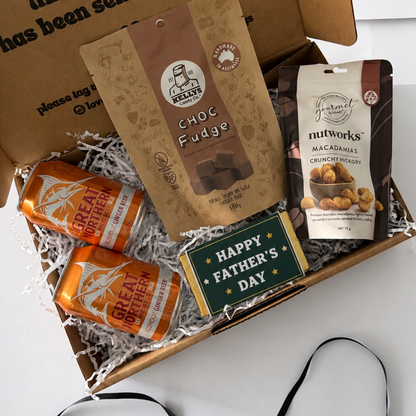 A Father's Day gift box set containing a bag of Choc Fudge, Nutworks Macadamia Crunchy Caramel, two orange cans of Great Britain, and a 'Happy Father's Day' card, arranged in shredded paper inside a brown cardboard box with a black ribbon.