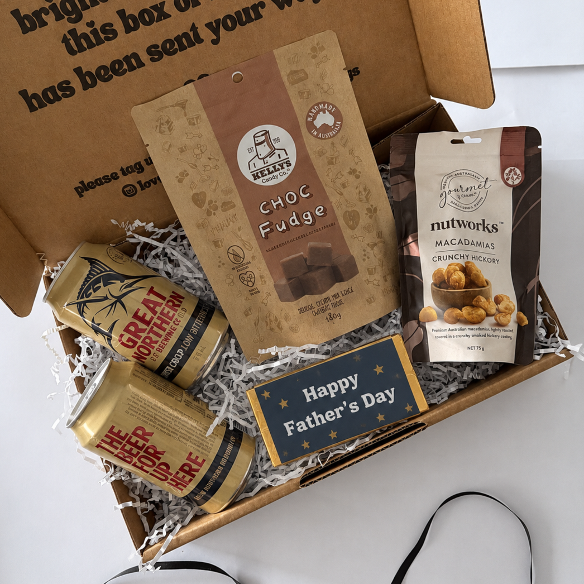 An open cardboard gift box filled with Father's Day treats and snacks, photographed from above on a white surface. The box contains a bag of Choc Fudge, a bag of Nutworks macadamia chips, two cans of Great Northern beans, and a small 'Happy Father's Day' card.