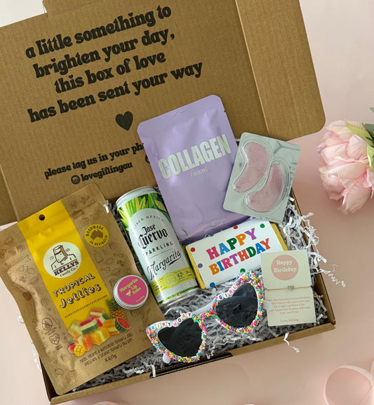 A curated gift box set displayed in an open brown cardboard box with printed text on the lid. Inside are a yellow bag of snacks, a green and white can, a lavender pouch labeled 'COLLAGEN', a sheet mask pack, a small card, novelty heart sunglasses, and a 'Happy Birthday' card.